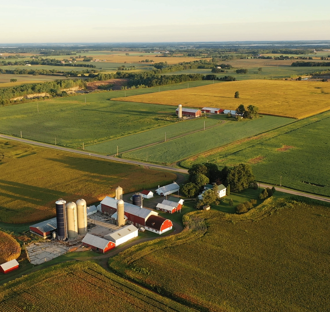 about h2 -2 Aerial view of a rural farm with multiple buildings, silos, and expansive green fields under a clear sky.